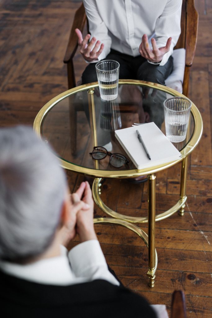 A therapist with gray hair sits in front of a table with a notebook across a talking client.