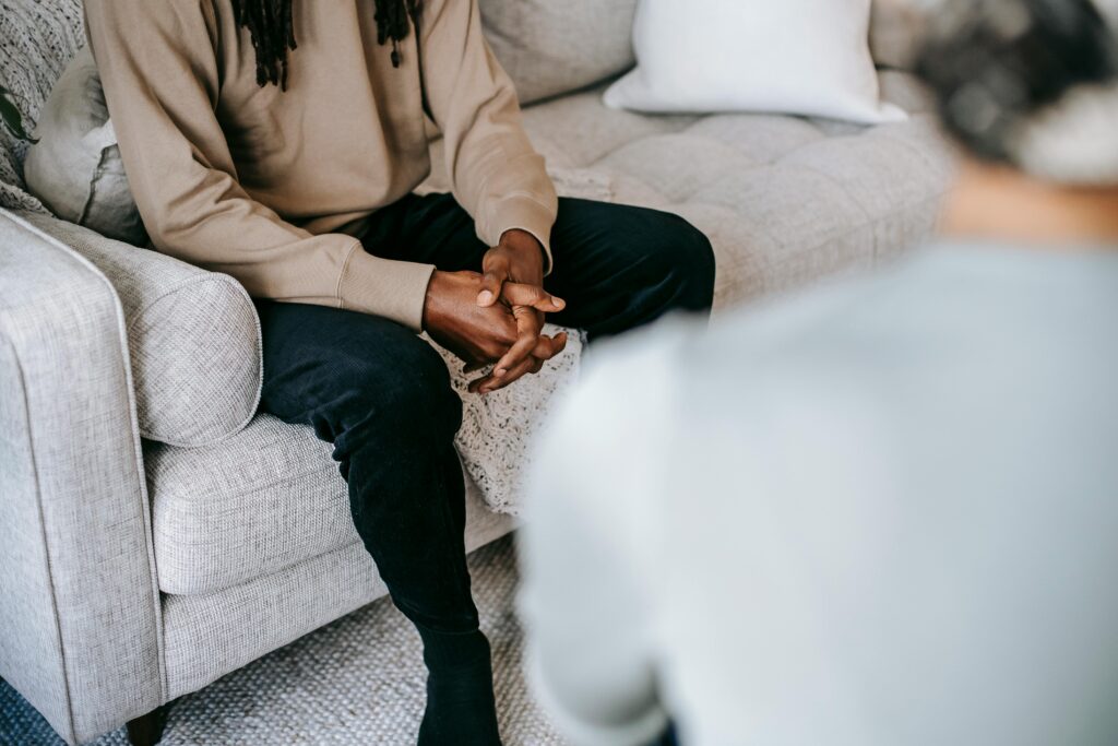 A black man sits on a couch while holding his hands together. Across from him is a therapist.