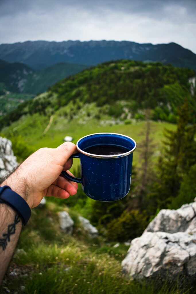 Left hand with a watch on the wrist holds a blue coffee mug in front of a grassy mountain background.